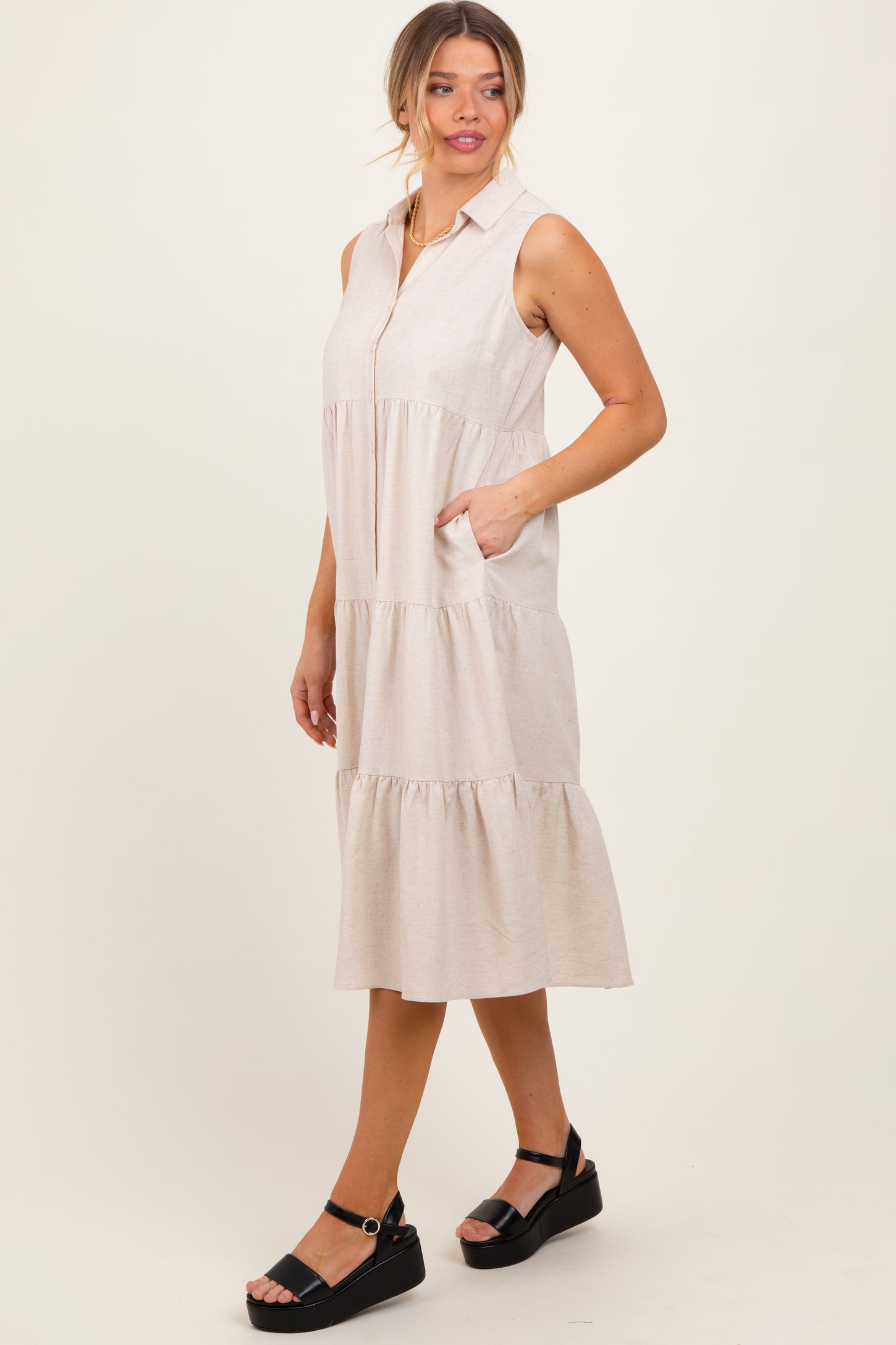woman wearing a light beige sleeveless collared midi dress with tiered hem and pockets, styled with black sandals