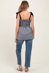 woman wearing a black and white gingham maternity top with adjustable straps and a cinched waist, paired with light blue jeans