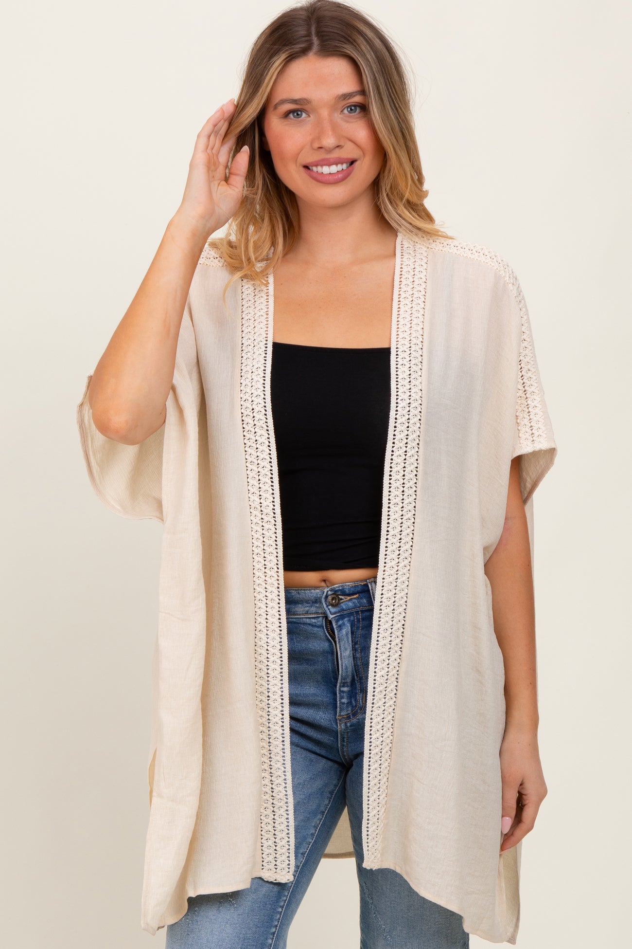 woman wearing a beige loose-fitting cardigan with lace details over a black crop top and blue jeans