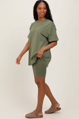 woman wearing an oversized olive green t-shirt and matching bike shorts, standing sideways with a relaxed pose