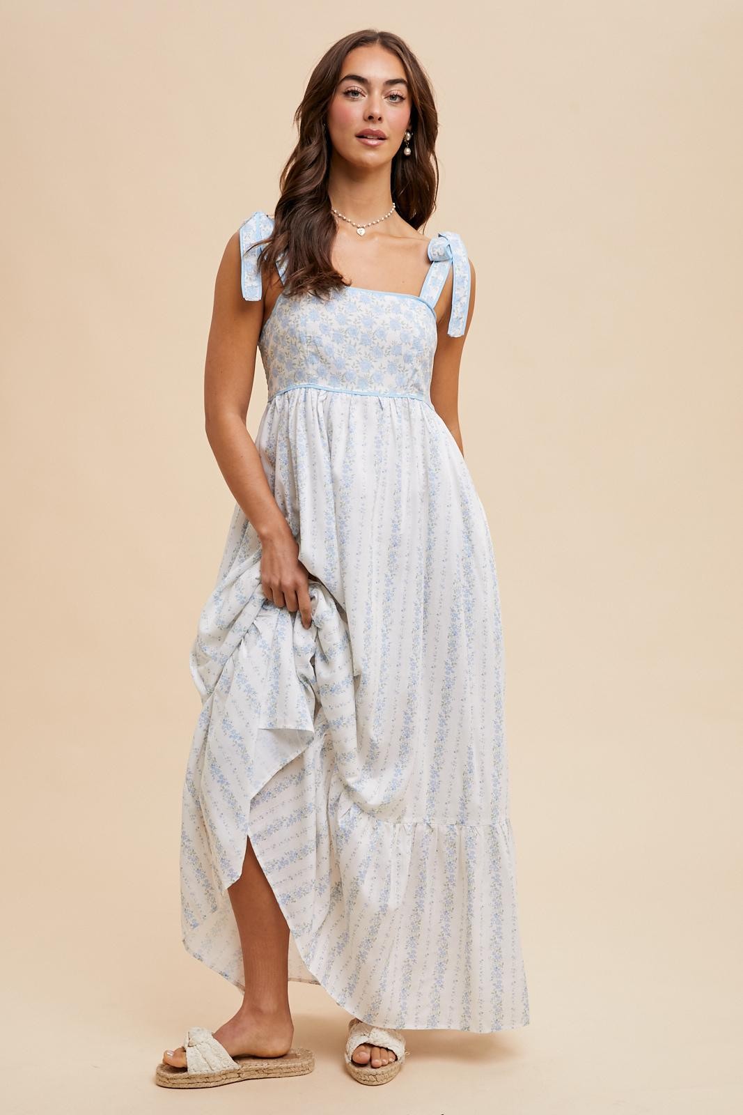 woman wearing a blue and white floral maxi dress with bow ties on the straps and a side slit, holding the dress in one hand