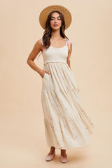 woman wearing a cream maxi dress with a smocked bodice, purple floral accents, pockets, and holding a straw hat