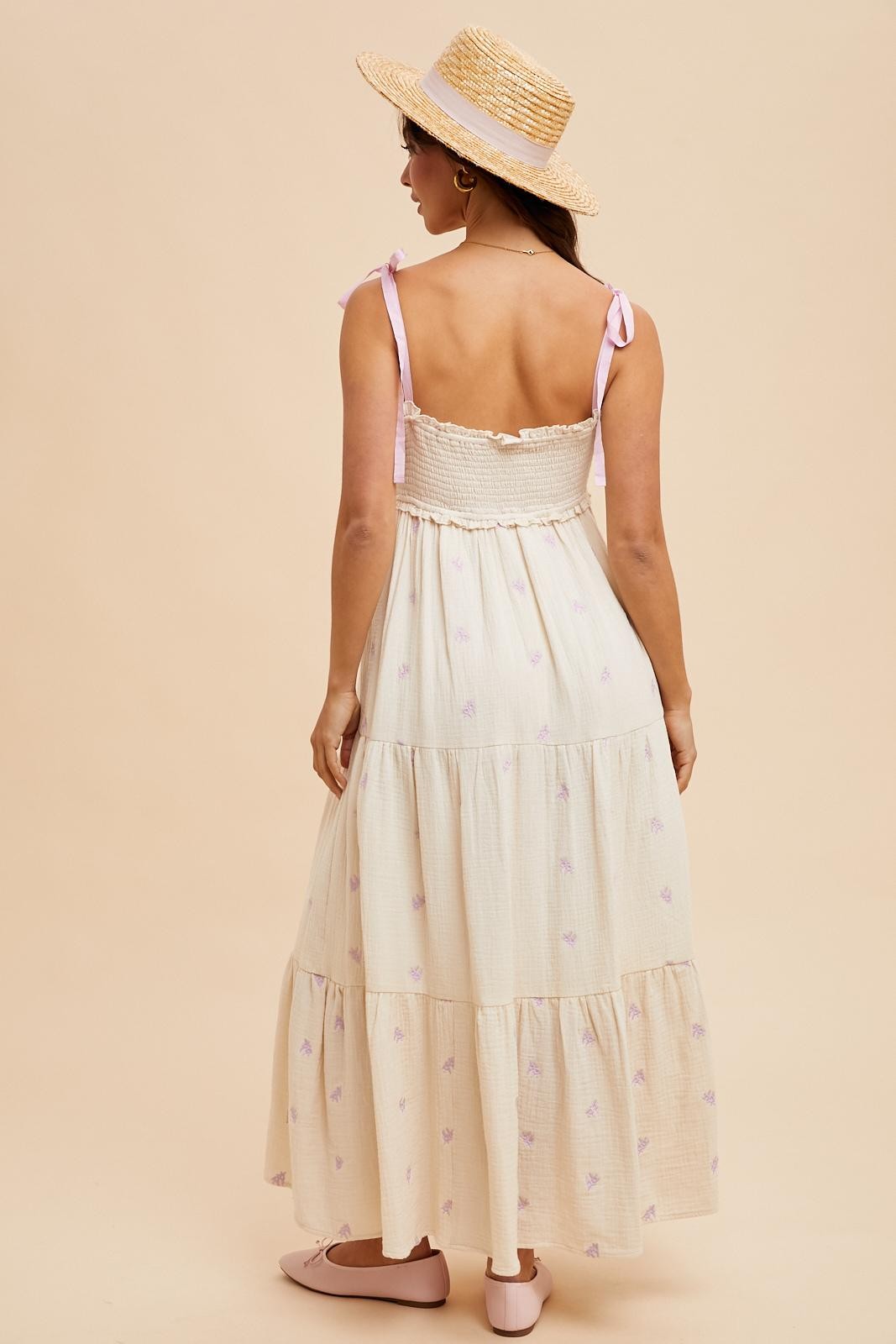woman wearing a cream tiered maxi dress with lilac floral details and adjustable straps, paired with a straw hat.