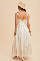 woman wearing a cream tiered maxi dress with lilac floral details and adjustable straps, paired with a straw hat.
