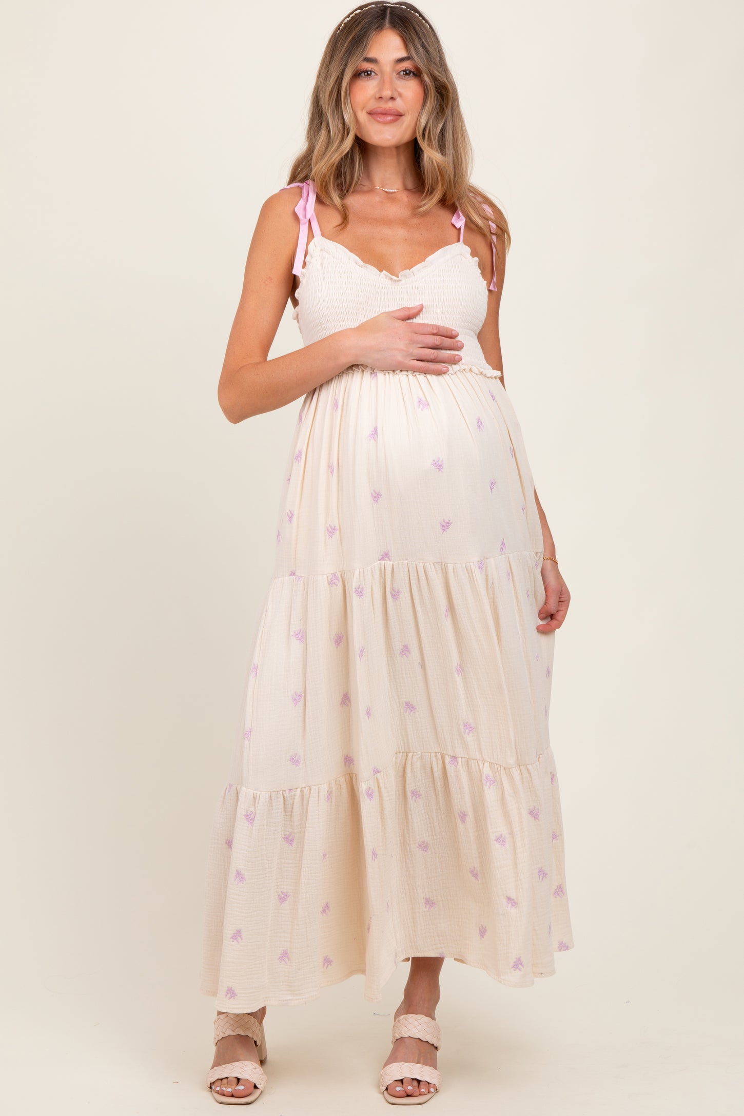 woman wearing a cream and pink floral maxi maternity dress, holding her baby bump and standing on a light background