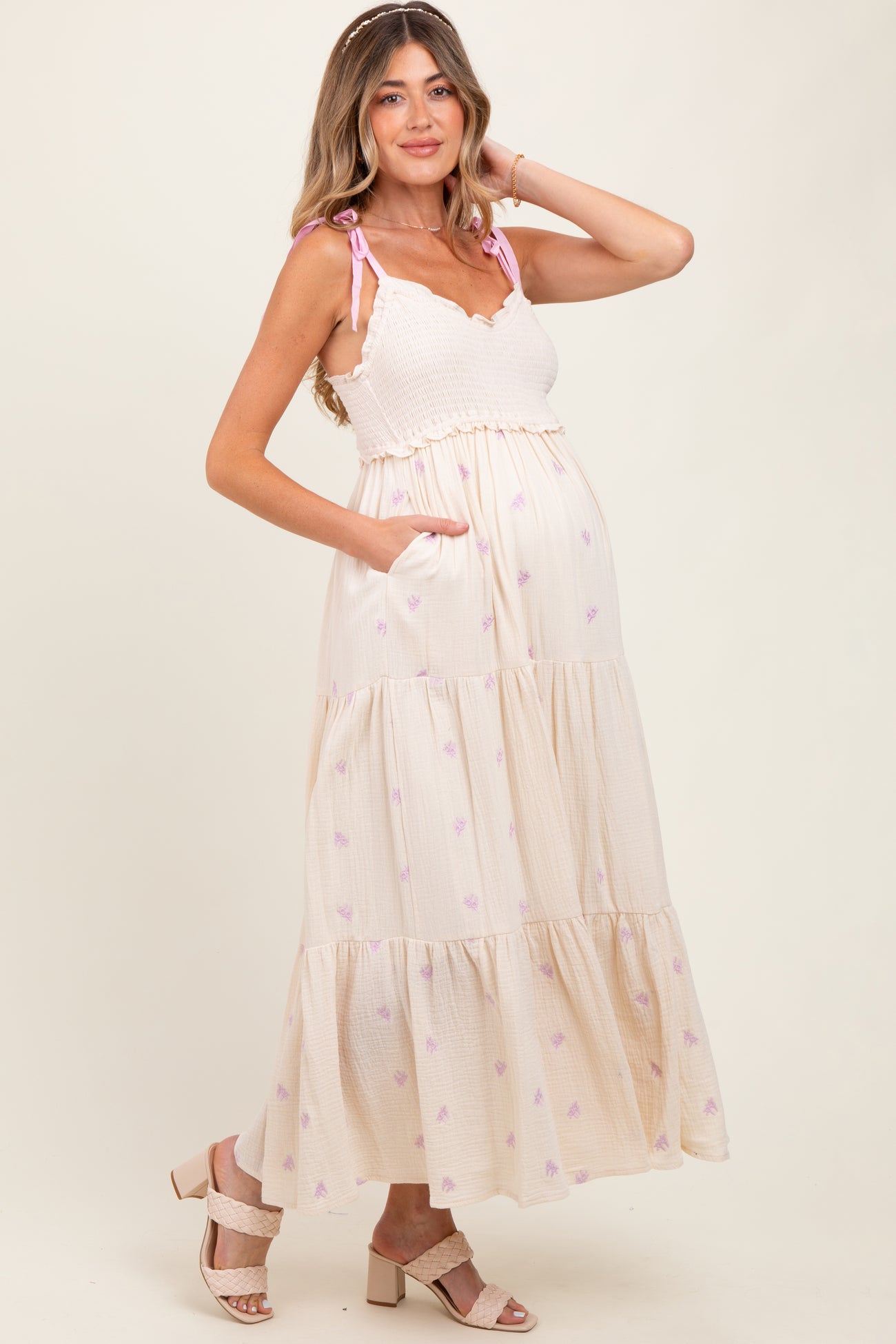 woman wearing a cream tiered maternity dress with pink floral details and adjustable straps, showcasing her baby bump