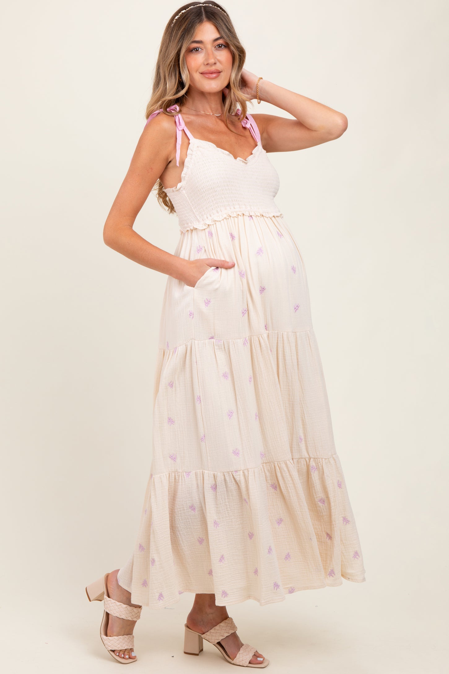 woman wearing a cream tiered maternity dress with pink floral details and adjustable straps, showcasing her baby bump