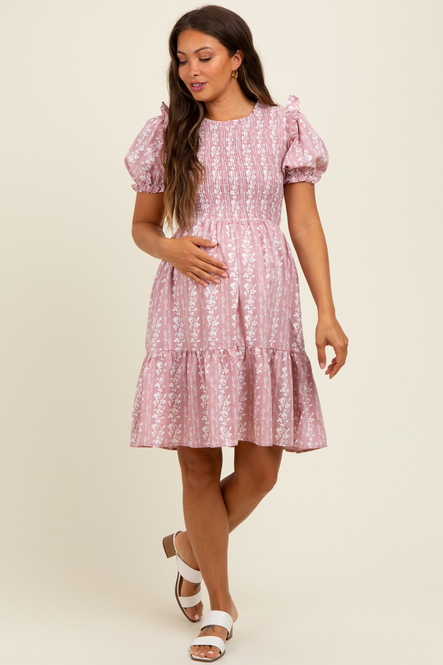 woman wearing a pink floral print smocked maternity dress with puff sleeves and a tiered skirt, holding her baby bump