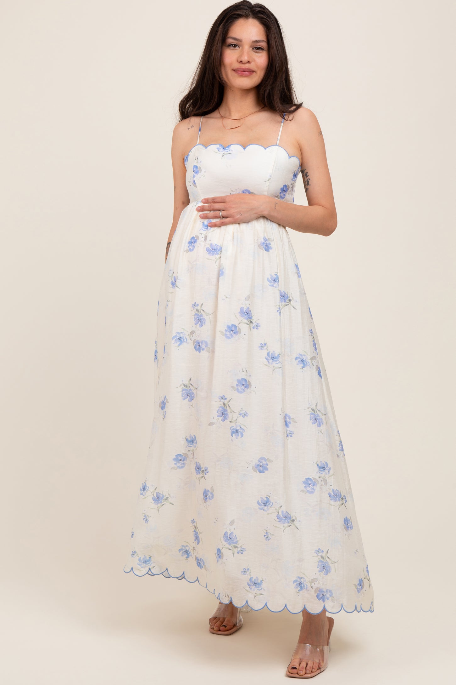 woman wearing a white floral maternity dress with scalloped edges and holding her baby bump