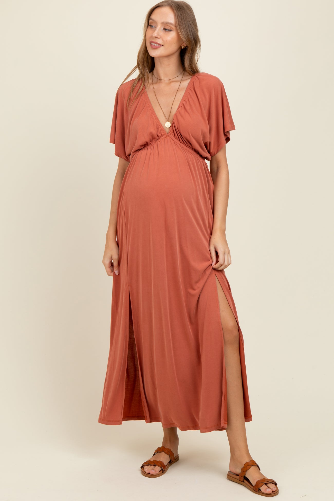 woman wearing a rust-colored maxi maternity dress with a deep V-neck and thigh-high split, showcasing her baby bump