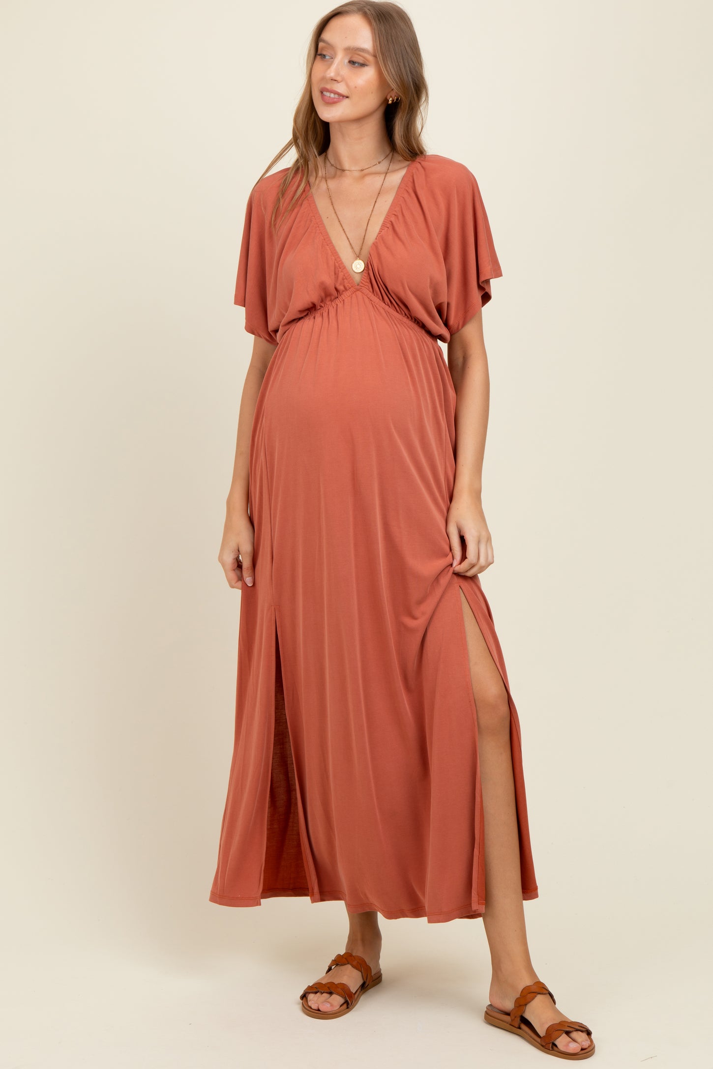 woman wearing a rust-colored maxi maternity dress with a deep V-neck and thigh-high split, showcasing her baby bump