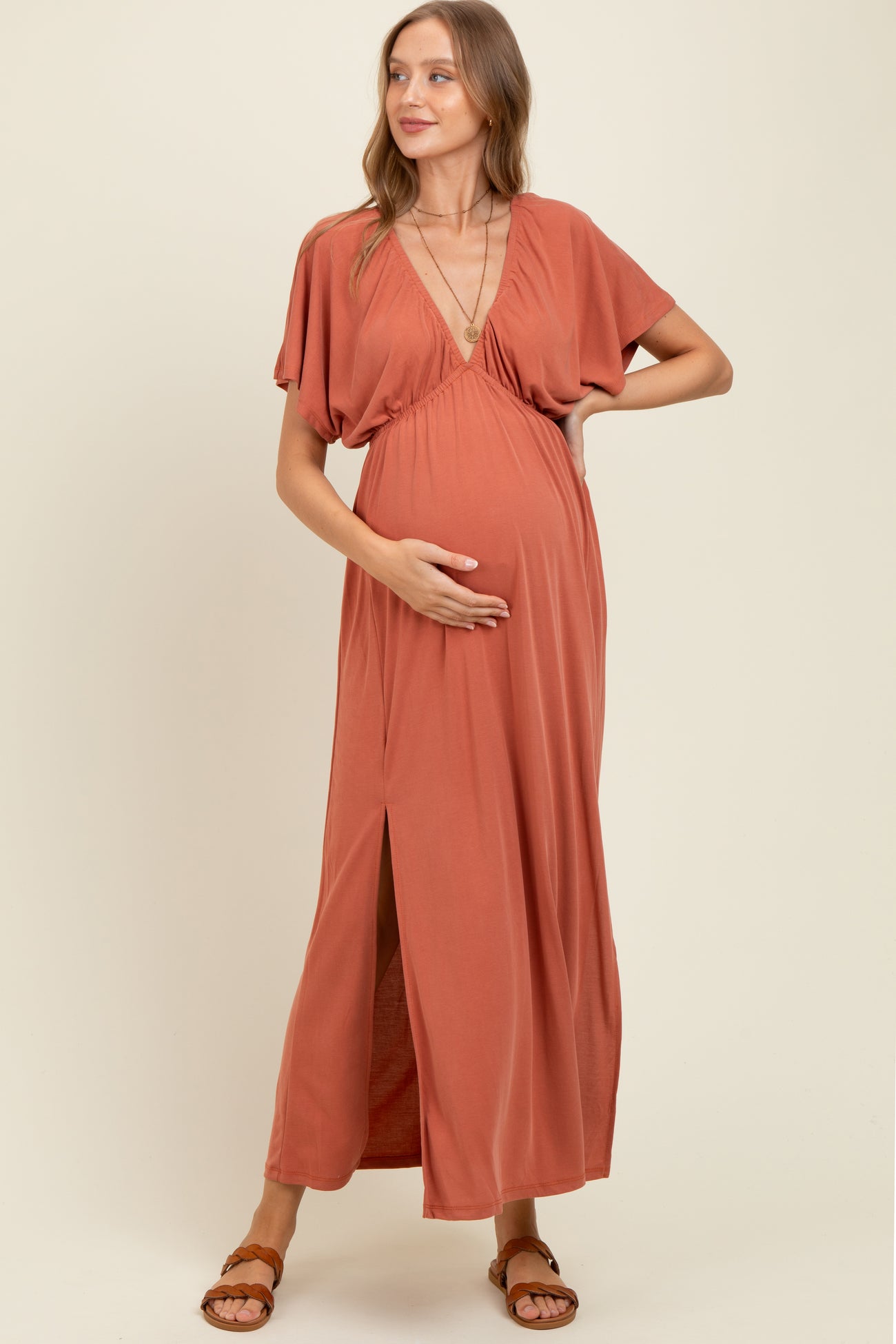 woman wearing a rust-colored maternity maxi dress with a deep V-neck and side slit, gently holding her baby bump