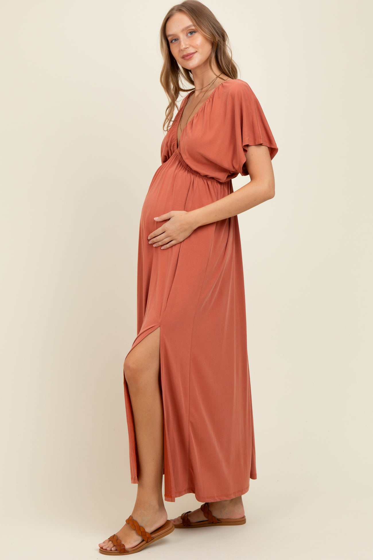 woman wearing a rust colored maternity maxi dress with short sleeves and a high slit, cradling her baby bump