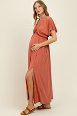 woman wearing a rust colored maternity maxi dress with short sleeves and a high slit, cradling her baby bump