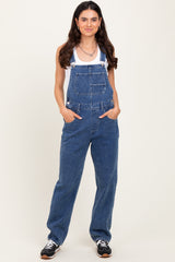 woman wearing a light blue denim overall with a white tank top underneath and hands in pockets, standing against a plain background