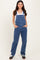 woman wearing a light blue denim overall with a white tank top underneath and hands in pockets, standing against a plain background
