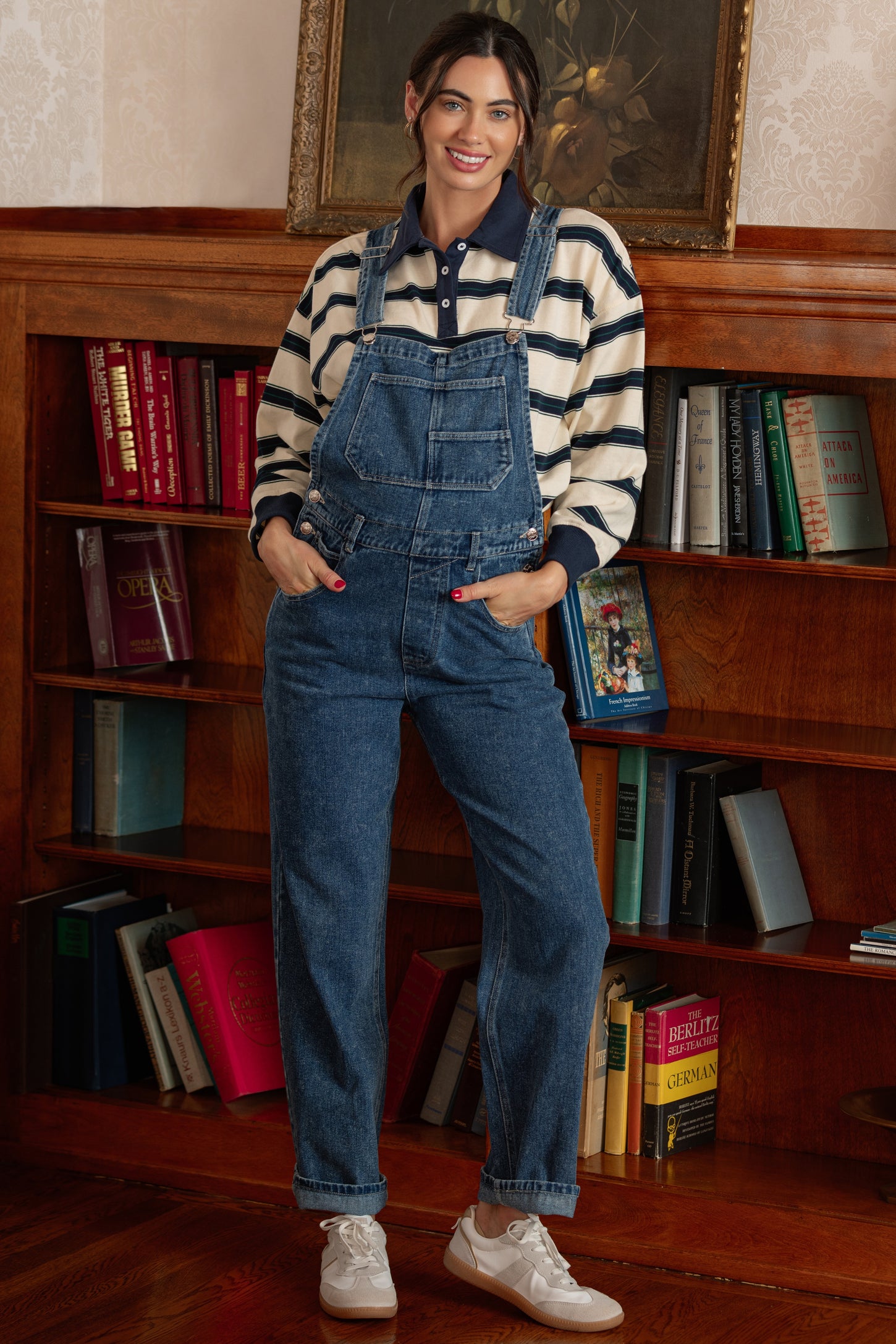woman wearing a striped long sleeve shirt and denim overalls with pockets, standing in a room with bookshelves