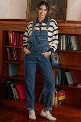 woman wearing a striped long sleeve shirt and denim overalls with pockets, standing in a room with bookshelves