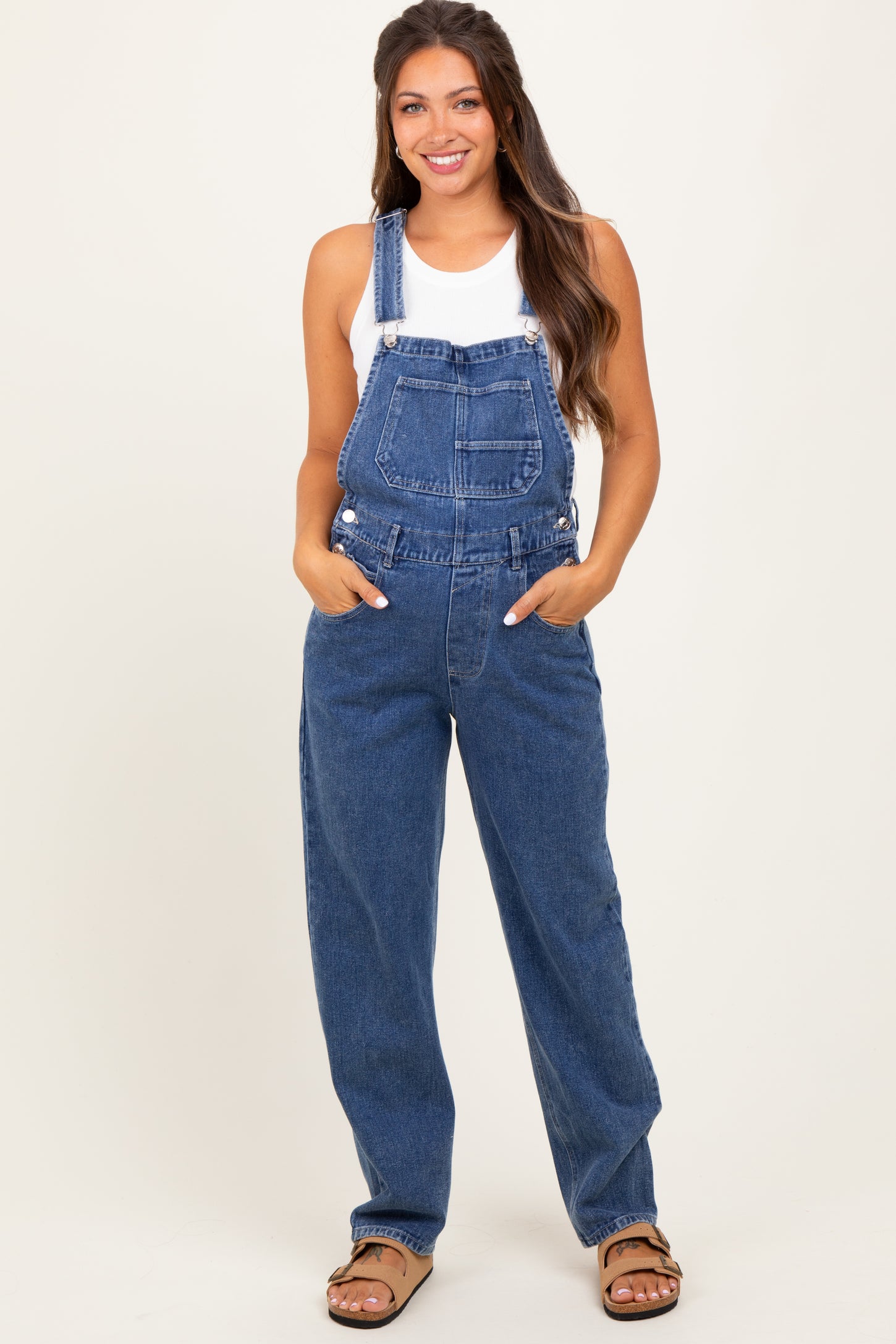 woman wearing a blue denim maternity overall with pockets and a white tank top, posing confidently