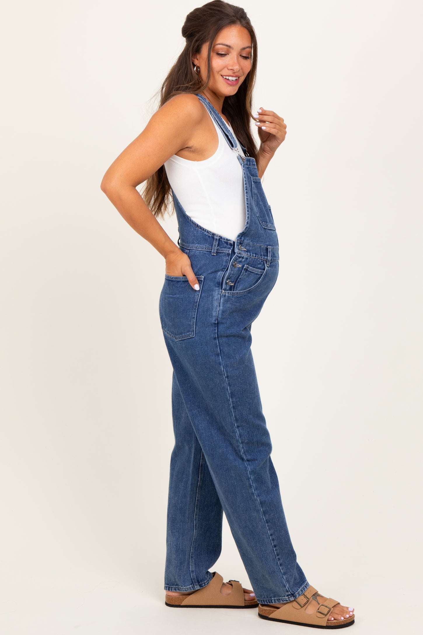 woman wearing a dark blue denim maternity overall with a white tank top and a visible baby bump