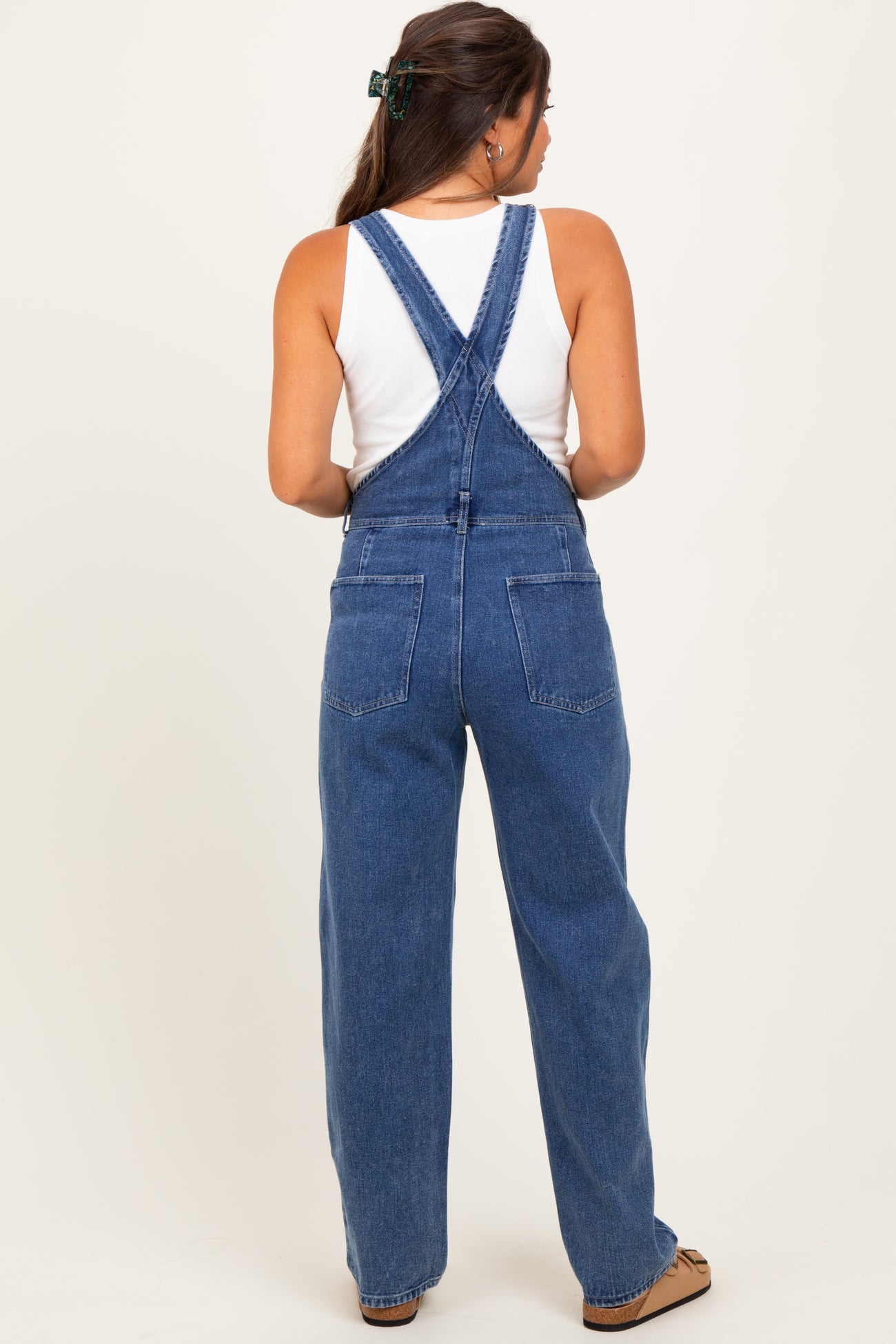 woman wearing a white tank top and blue denim maternity overalls with wide legs, showing her back