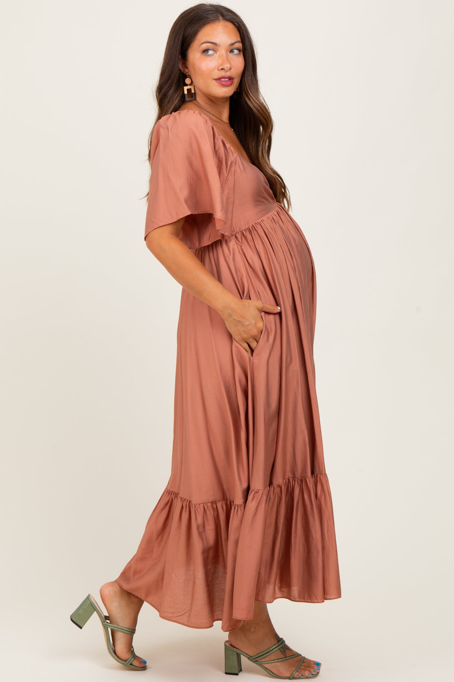woman wearing a rust-colored maternity maxi dress with short sleeves and a tiered ruffle hem, showcasing her baby bump