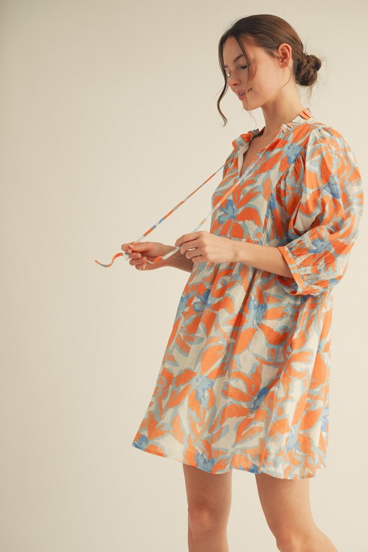 woman wearing a light beige floral print dress with orange and blue leaves and puff sleeves, holding a tie at the neckline