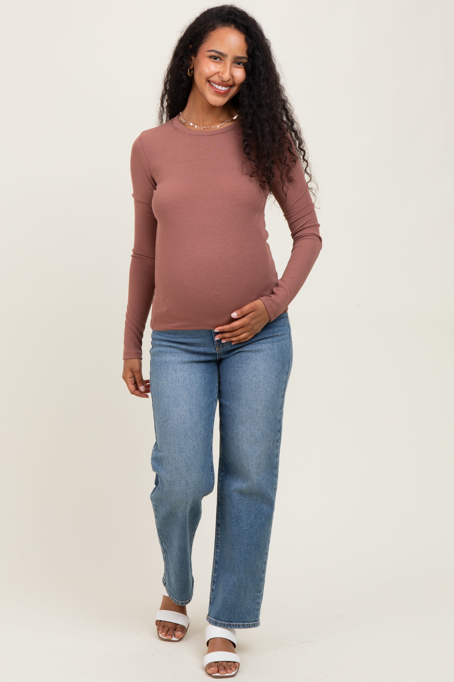 woman wearing a rust ribbed long sleeve maternity top and blue straight leg jeans, holding her baby bump
