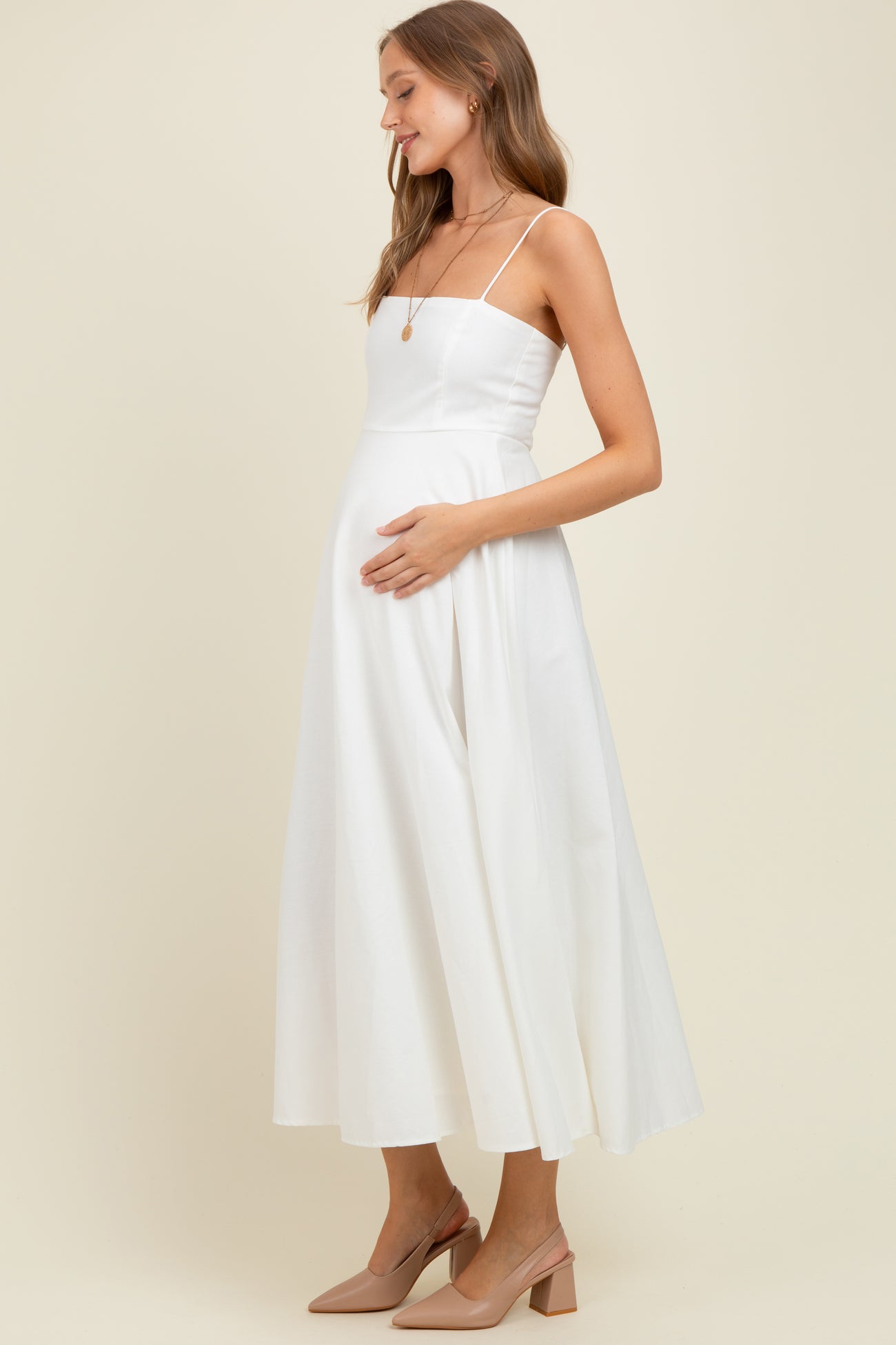 woman wearing a white maternity dress with spaghetti straps and a flowing skirt, holding her baby bump