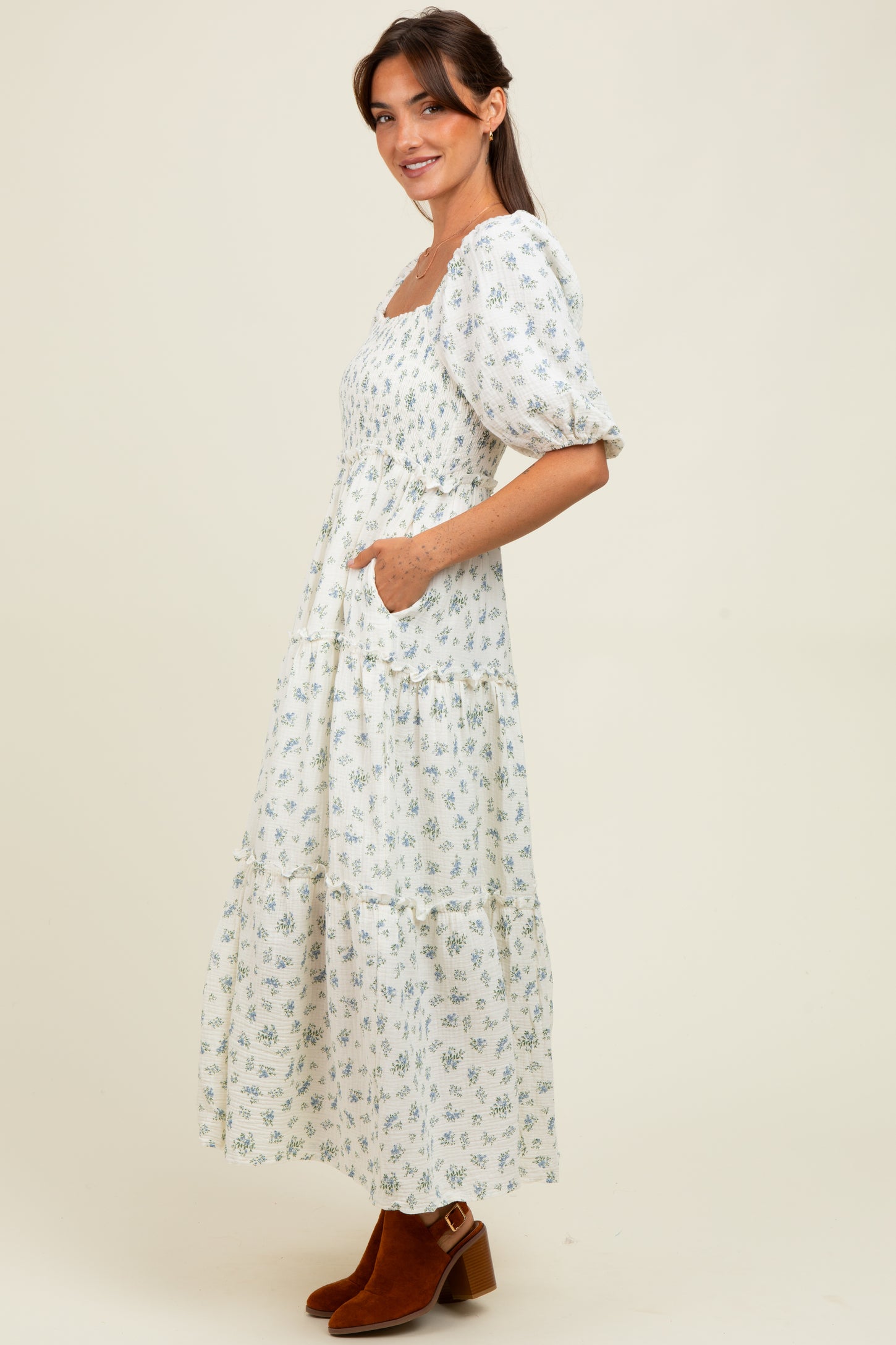 woman wearing a white floral maxi dress with puff sleeves and pockets, standing in profile with brown ankle boots