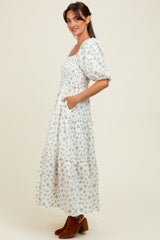 woman wearing a white floral maxi dress with puff sleeves and pockets, standing in profile with brown ankle boots