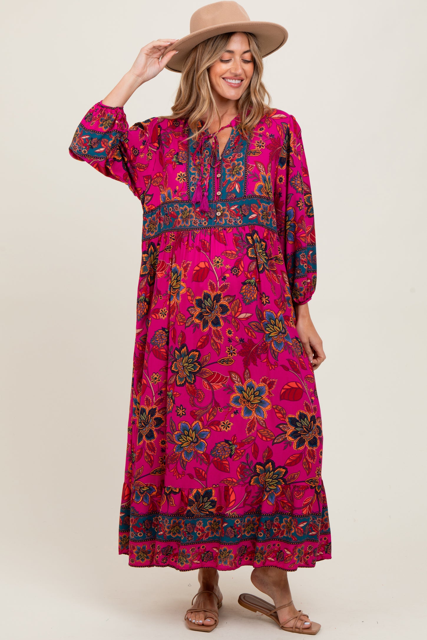 woman wearing a pink floral maternity maxi dress with long sleeves and a tiered hem, styled with a hat and sandals