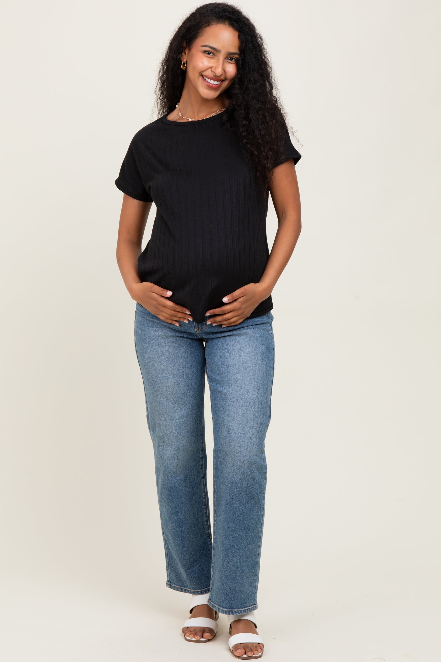 woman wearing a black ribbed maternity top and blue jeans, gently cradling her baby bump
