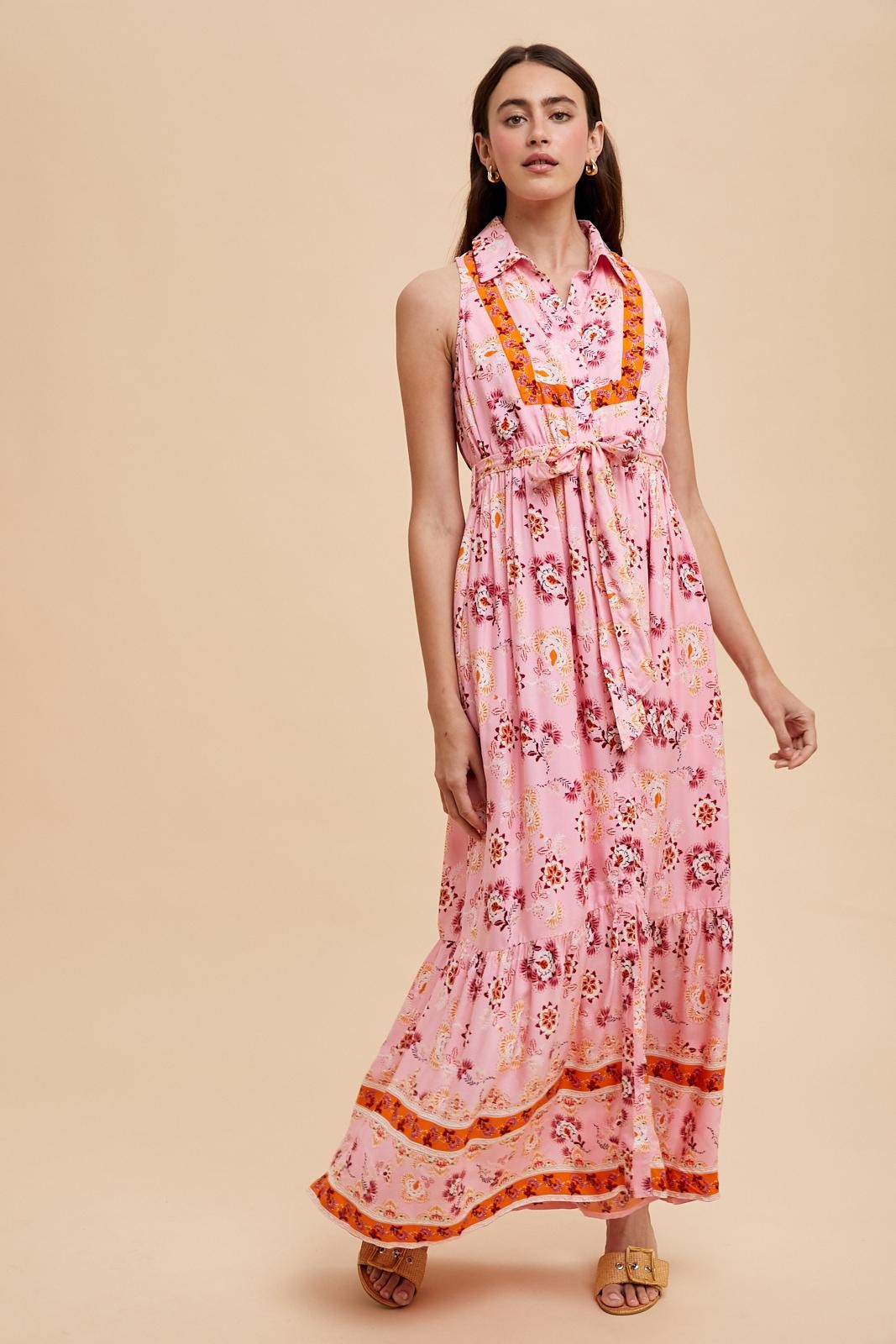 woman wearing a pink floral maxi dress with a tie at the waist and orange accents, posing against a light background