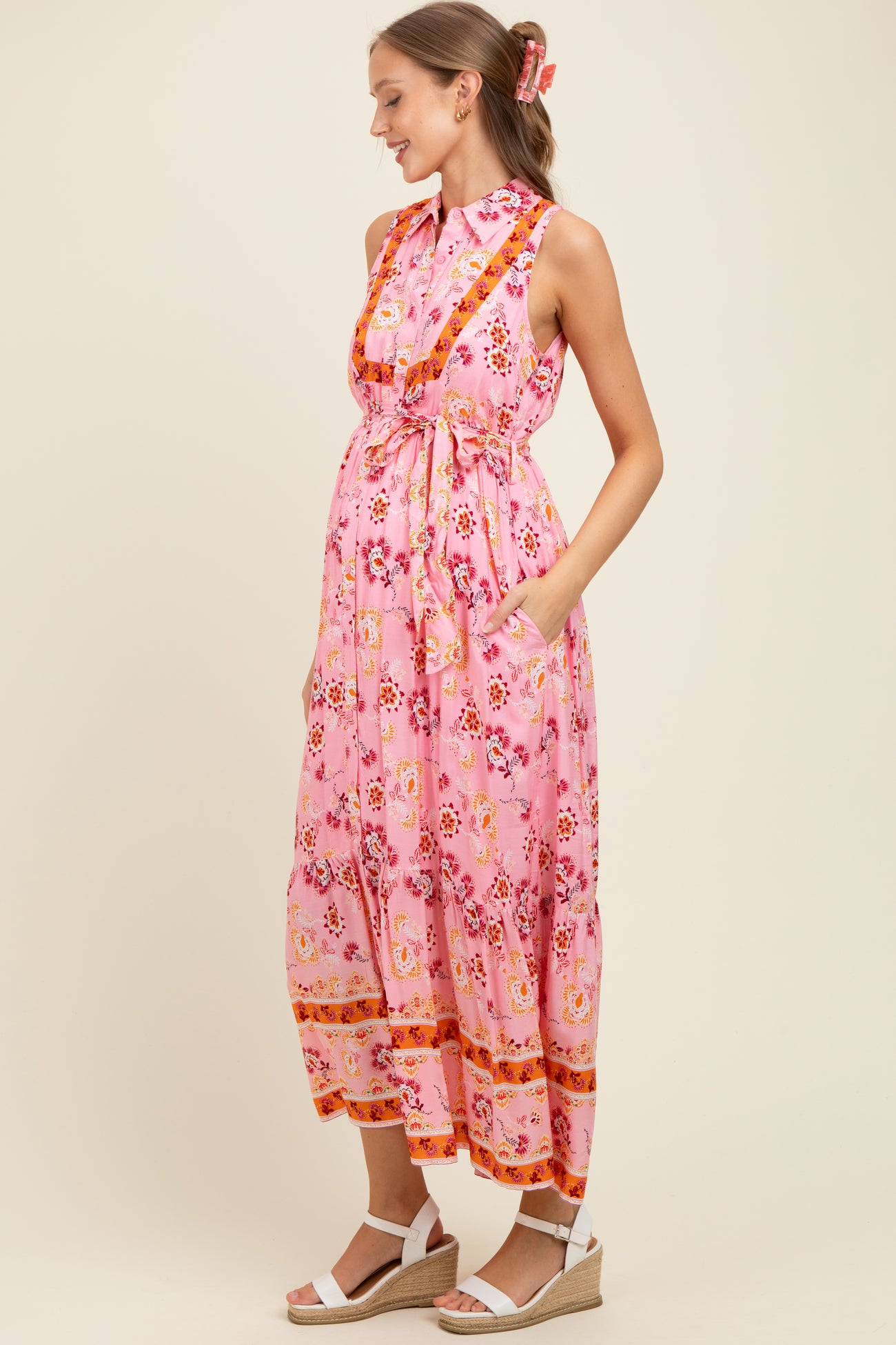 woman wearing a pink floral maternity dress with a collared neck, belted waist, and pockets, showcasing her baby bump