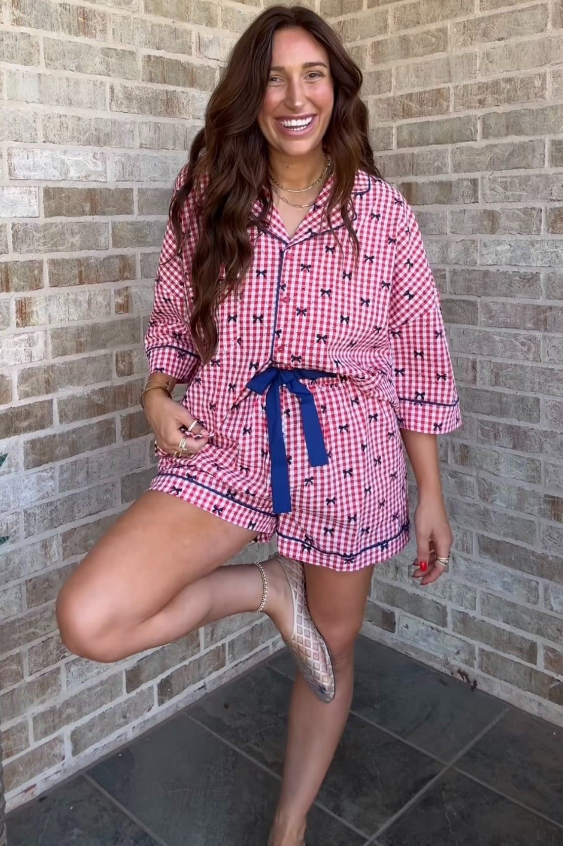 woman wearing a pink and white checkered short pajama set with a navy tie belt, smiling while standing on one leg
