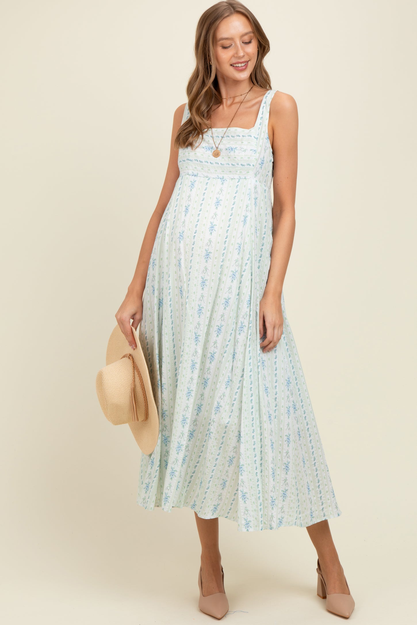 woman wearing a light blue floral maternity dress with a fitted bodice and flowing skirt, holding a straw hat and showing her baby bump