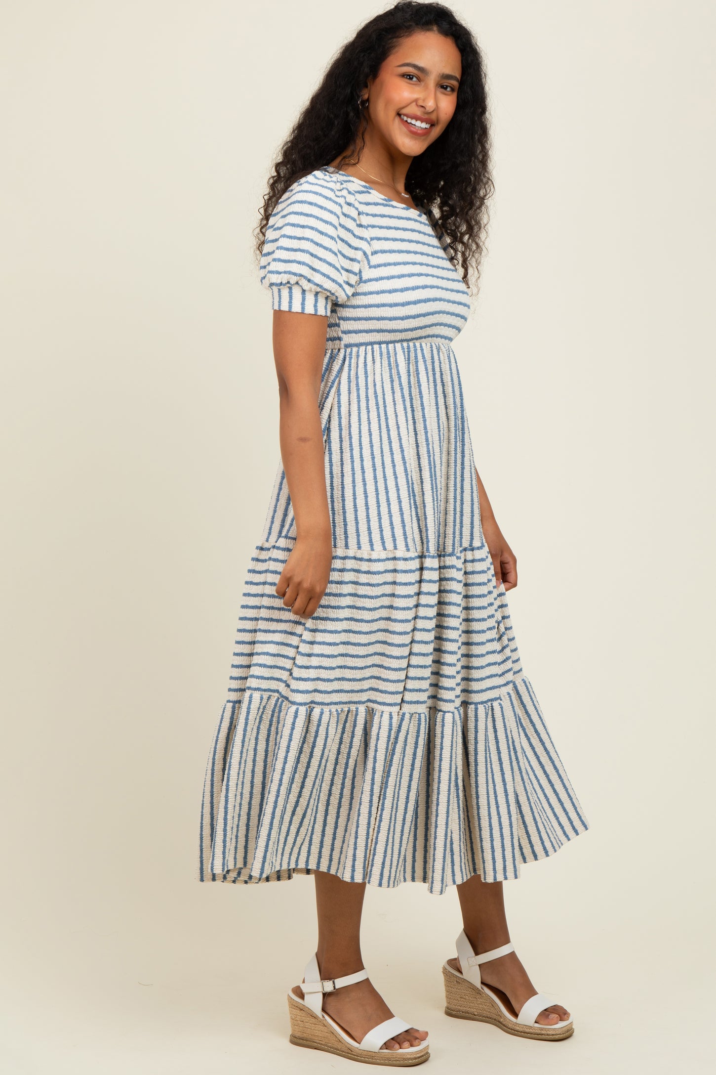 woman wearing a striped blue and white maxi dress with short puff sleeves and tiered skirt, standing in a neutral background
