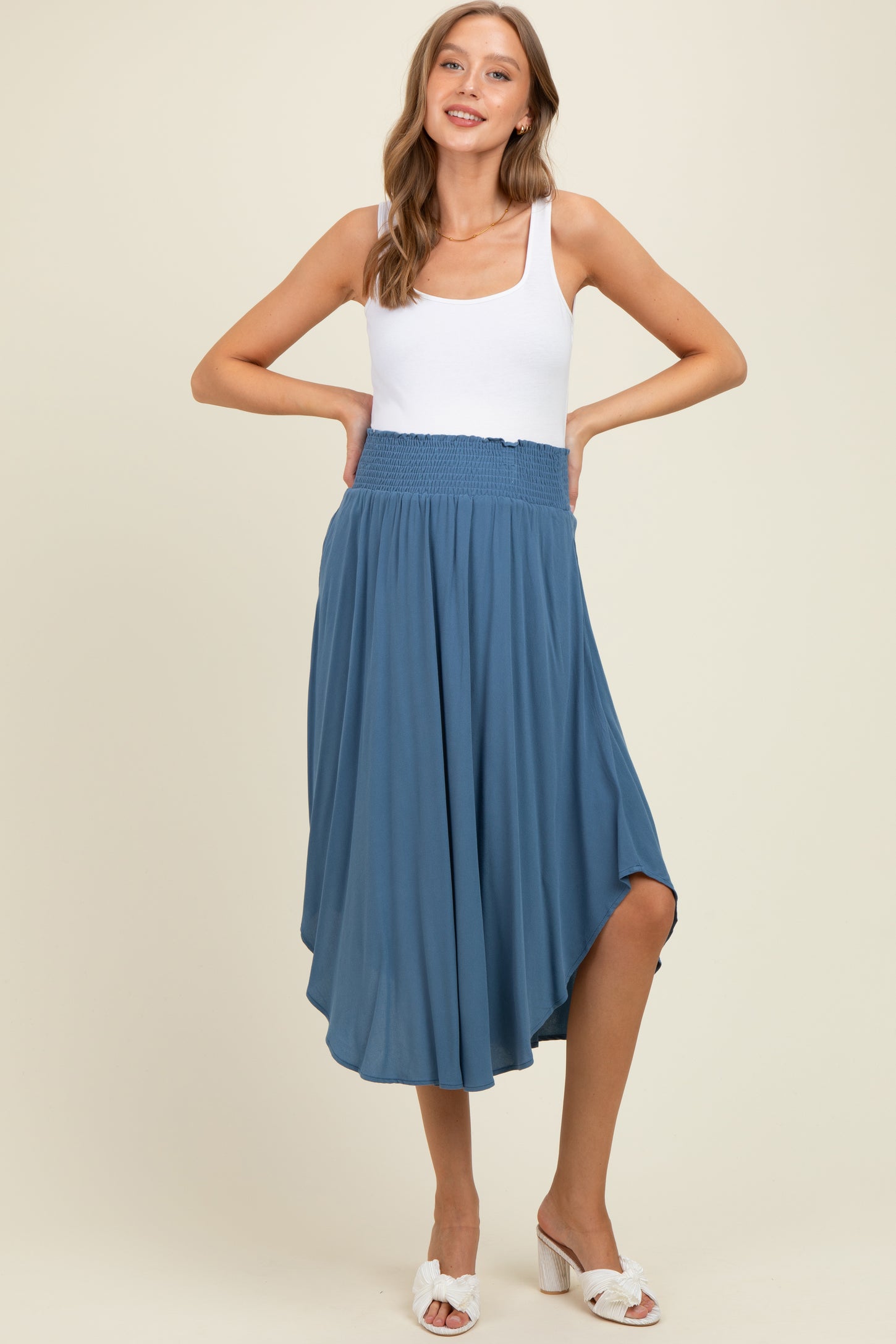 woman wearing a white tank top and a flowy blue maternity maxi skirt, posing with hands on hips