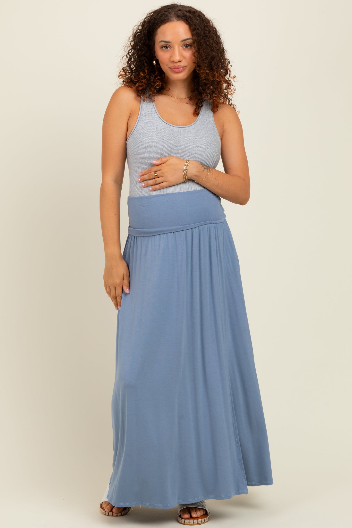 woman wearing a light gray tank top and a flowy blue maternity maxi skirt, gently holding her baby bump