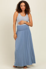 woman wearing a light gray tank top and a flowy blue maternity maxi skirt, gently holding her baby bump