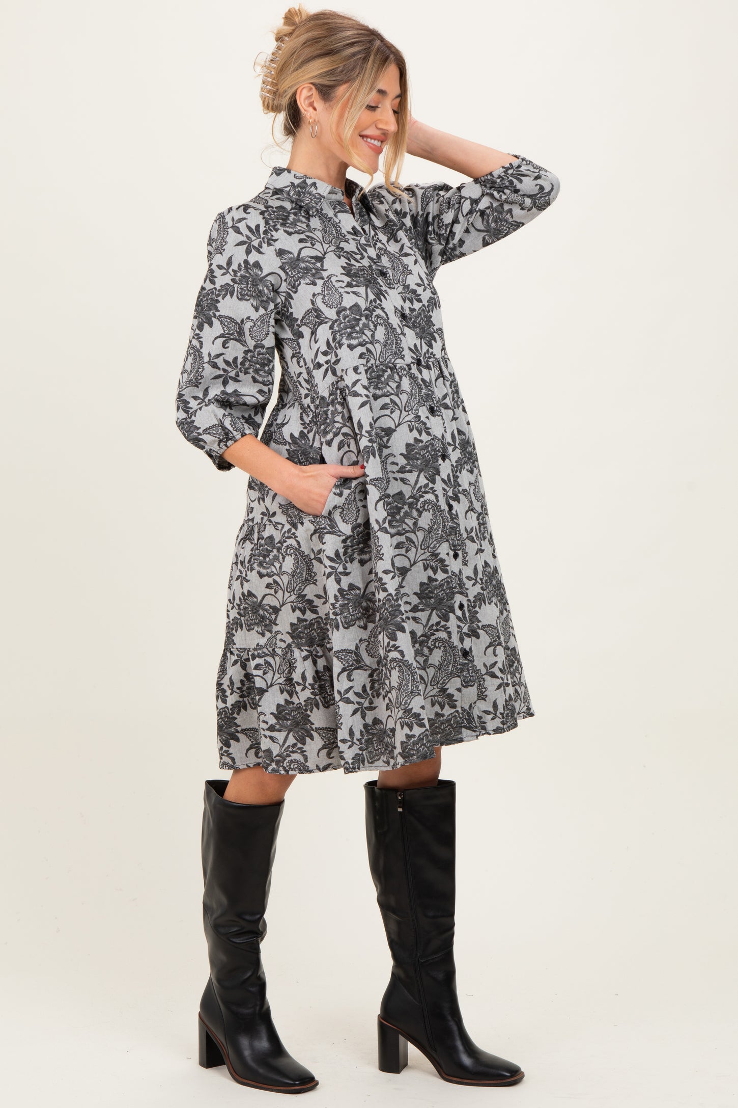 woman wearing a black and gray floral maternity dress with pockets and knee-high black boots, showing her baby bump