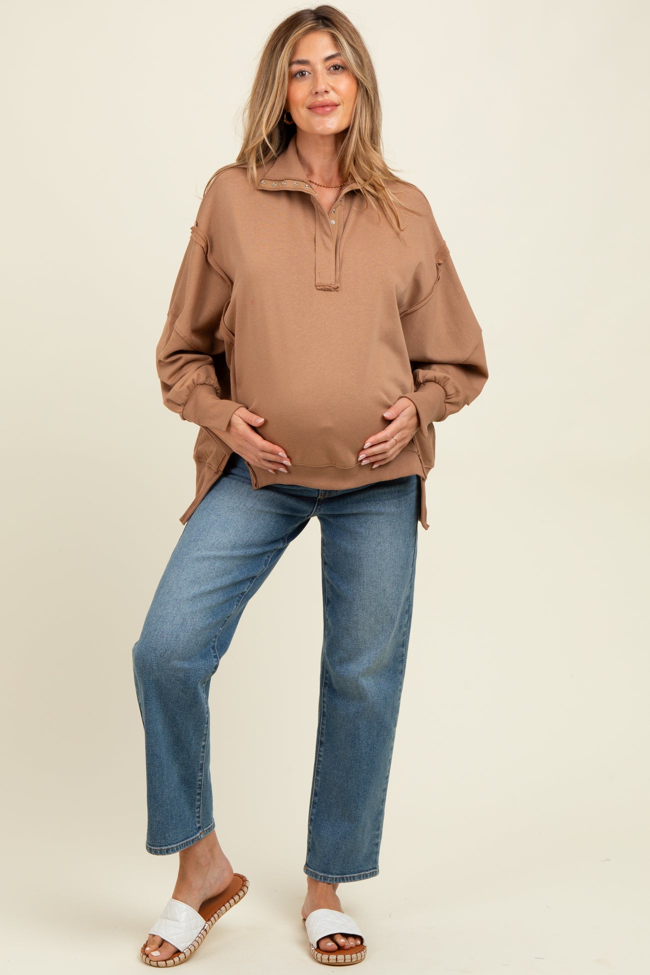 woman wearing a brown maternity pullover and blue jeans, cradling her baby bump