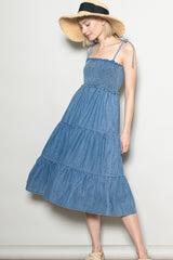 woman wearing a light denim tiered maxi dress with a smocked bodice and adjustable tie straps, styled with a straw hat