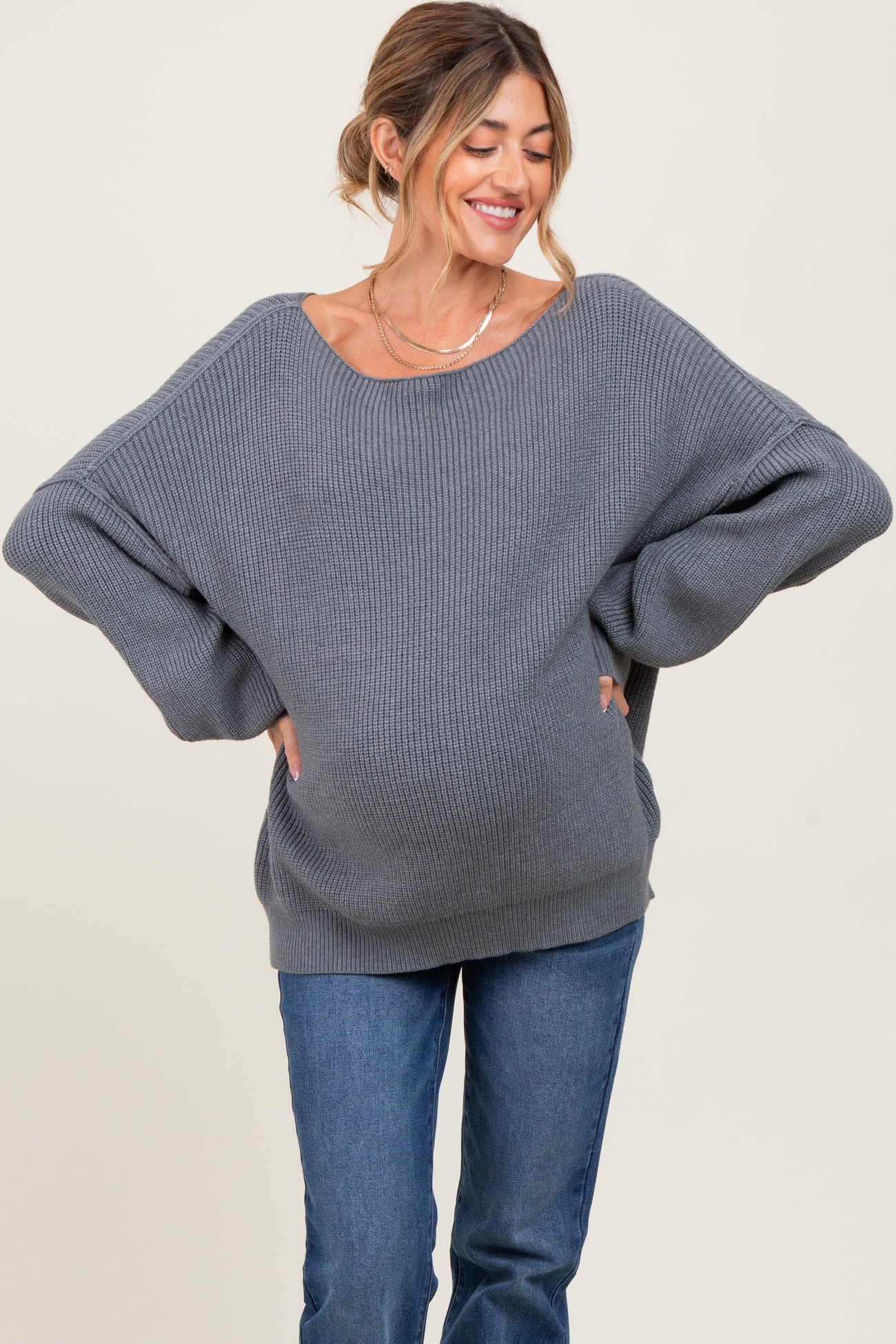 woman wearing a gray oversized ribbed maternity sweater with a visible baby bump and smiling while posing