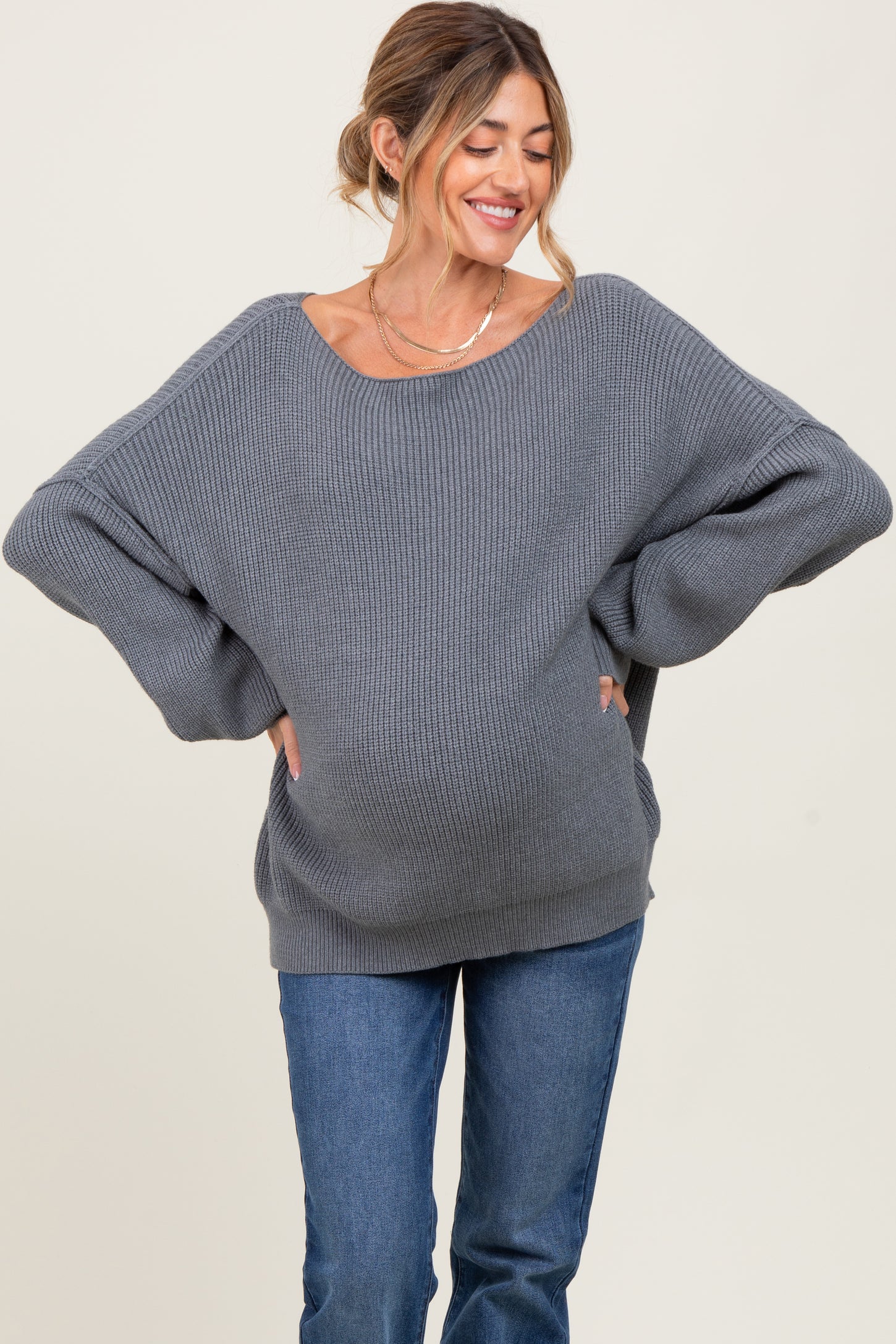 woman wearing a gray oversized ribbed maternity sweater with a visible baby bump and smiling while posing