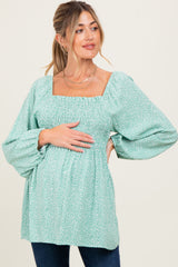 woman wearing a green floral maternity top with square neckline and puffed sleeves, gently resting her hands on her baby bump