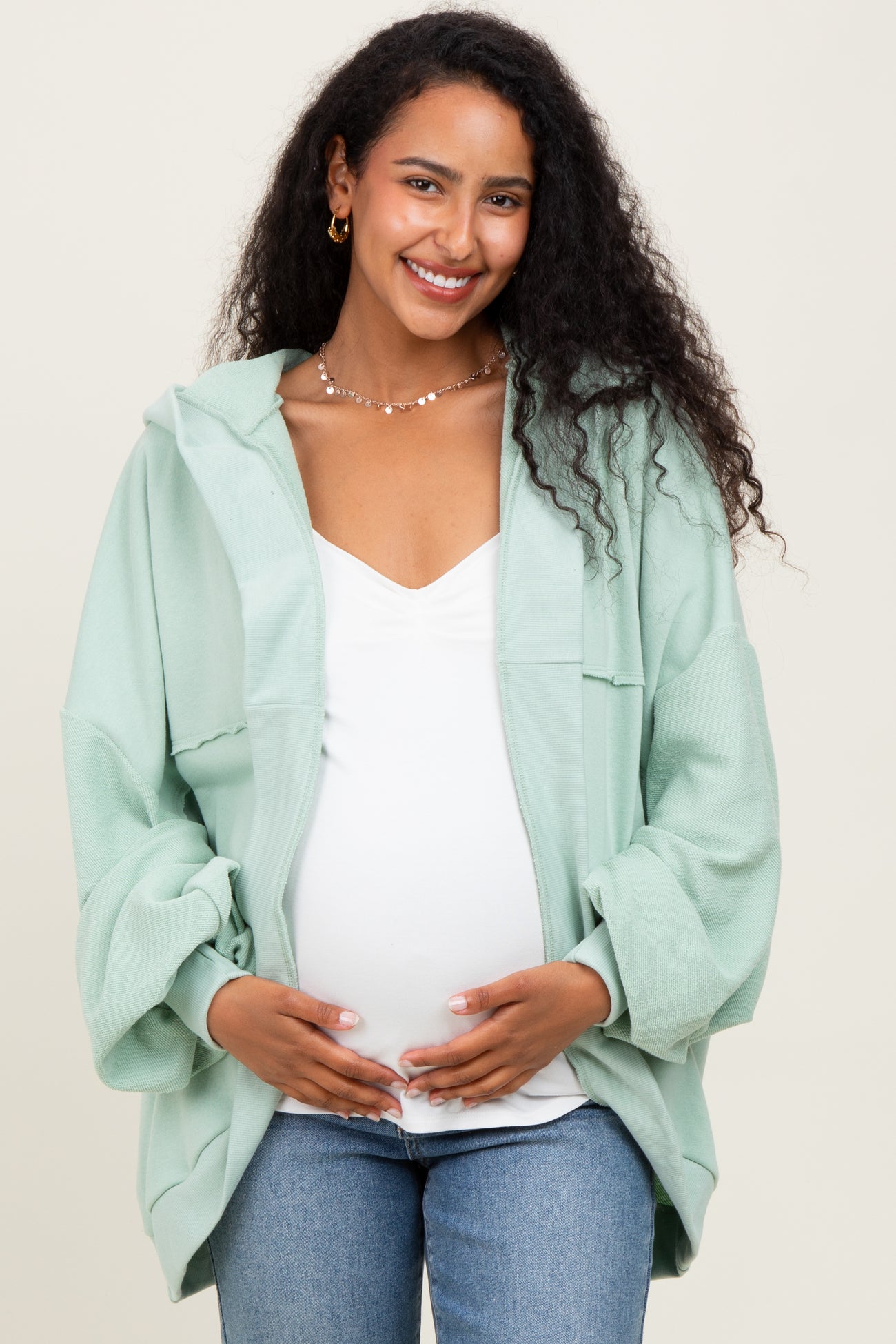 woman wearing a light green maternity zip-up hoodie over a white top and holding her baby bump