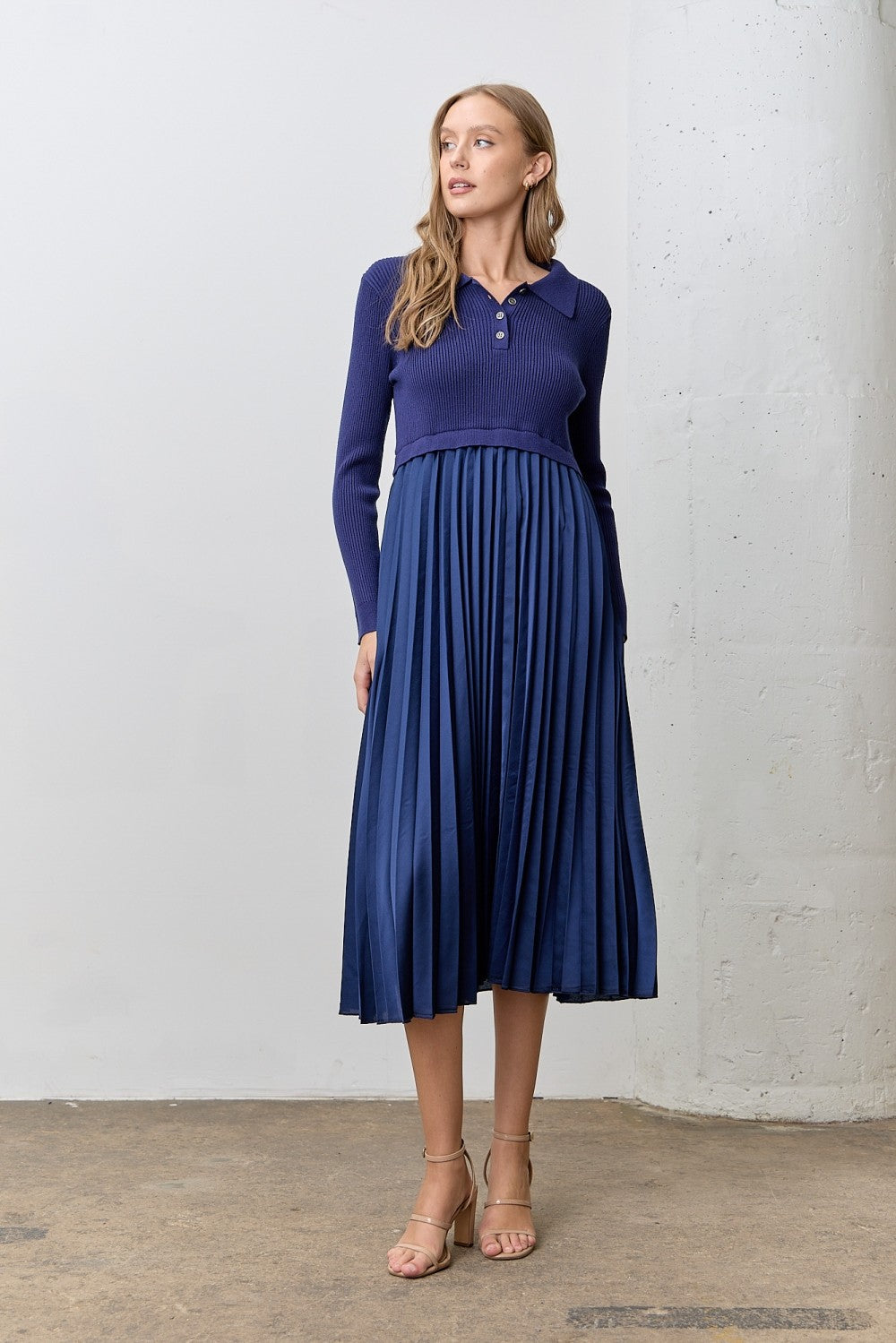 woman wearing a navy ribbed polo sweater and a pleated skirt dress, standing in a minimalist setting