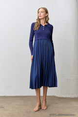 woman wearing a navy ribbed polo sweater and a pleated skirt dress, standing in a minimalist setting